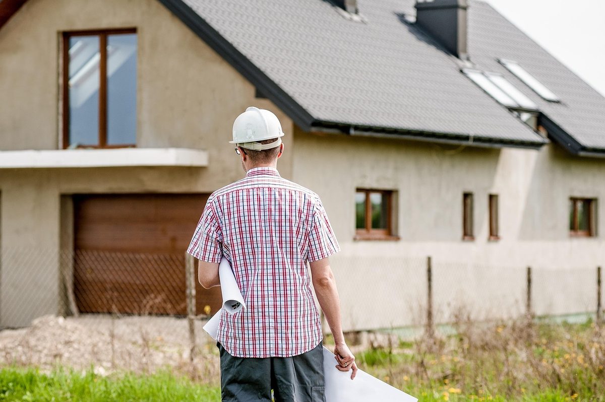 A contractor in a hard hat walks toward a house carrying rolled plans